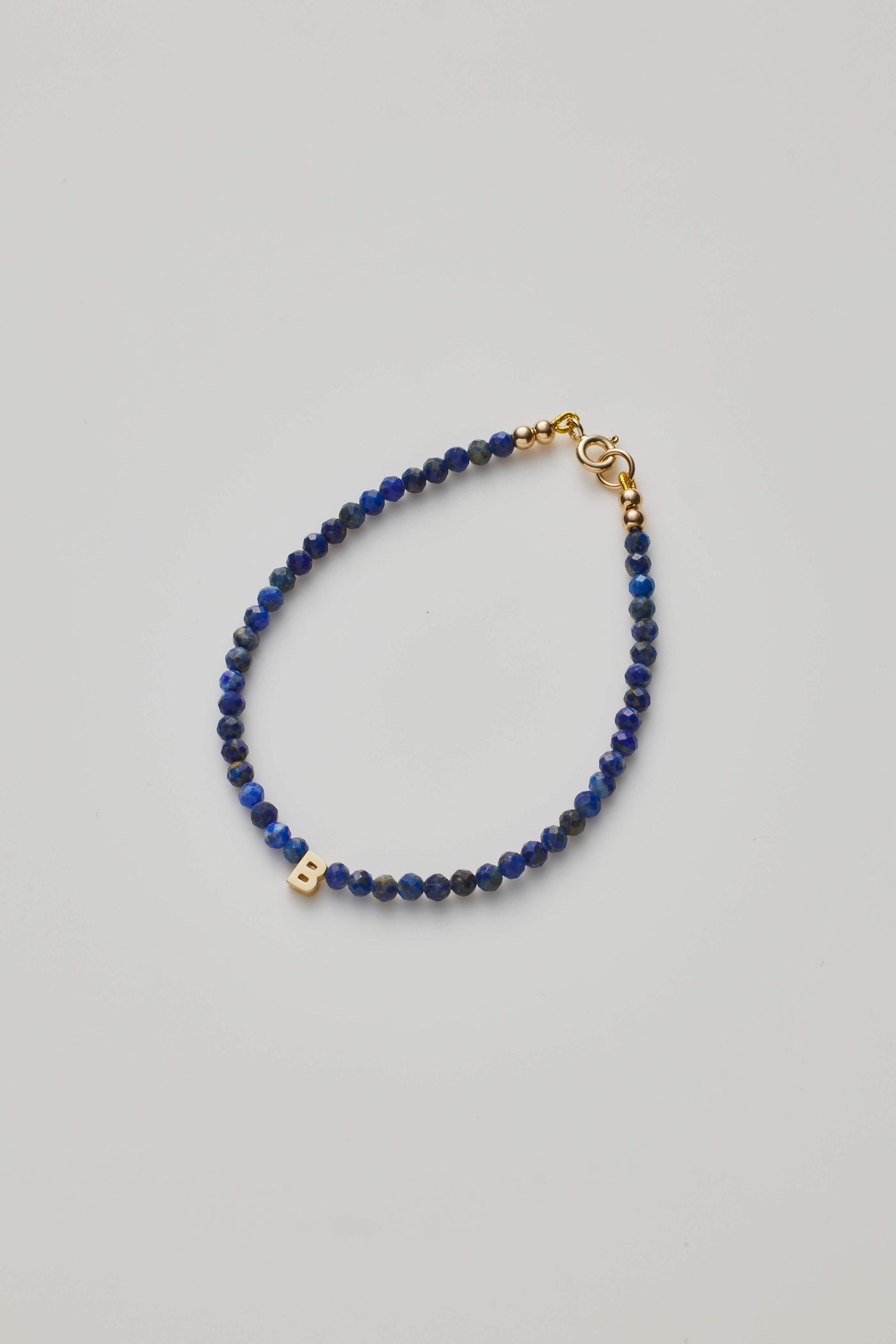 The One Letter Bracelet in Lapis Lazuli and 14k Gold is a delicate piece featuring small, round lapis lazuli beads and an 18k gold clasp. Among the hand-strung blue beads, a gold letter "B" charm is prominently placed, adding a personalised touch to this elegant bracelet, which is displayed on a plain white background.