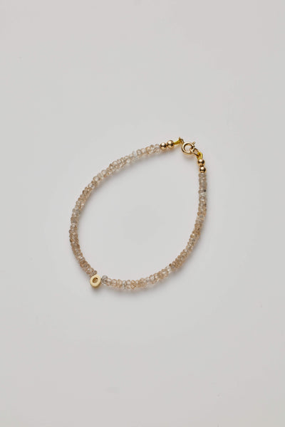 Introducing the One Letter Bracelet in Zircon and 14k Gold: a delicate bracelet adorned with a thin strand of small, translucent zircon beads. A single 14k solid gold bead adds a touch of sophistication, and the bracelet is secured with a gold clasp. The simple yet elegant design stands out beautifully against a plain, light background. Custom sizes are available upon request.