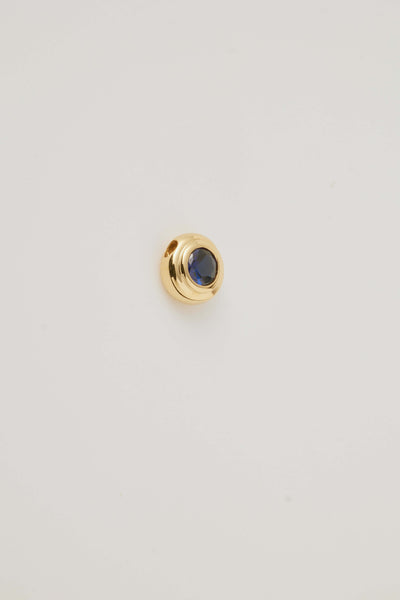 Side view image of the September Birthstone Charm featuring a deep blue sapphire gemstone in an elegant gold setting. Photographed on a light grey background.