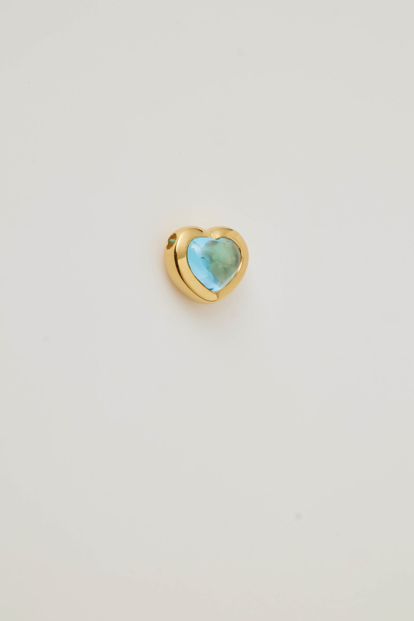 Side profile of a blue topaz cabochon stone set in a polished gold heart frame, displayed on a light grey background. Elegant and timeless.