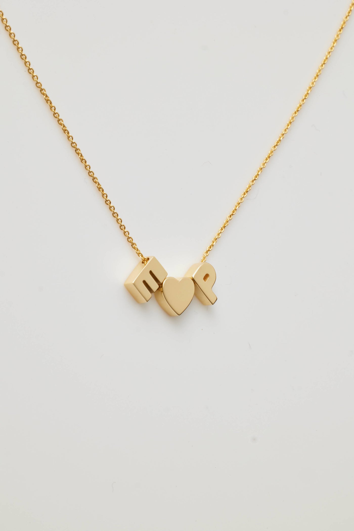 The Two Letters and Heart Necklace in 18k Gold boasts a delicate chain adorned with three exquisite charms: a capital letter "E" a heart symbol centrally placed, and a capital letter "P." Made from 18k solid gold, this beautiful piece is displayed against a simple, light-coloured backdrop and features an openable connector clasp for effortless wear.