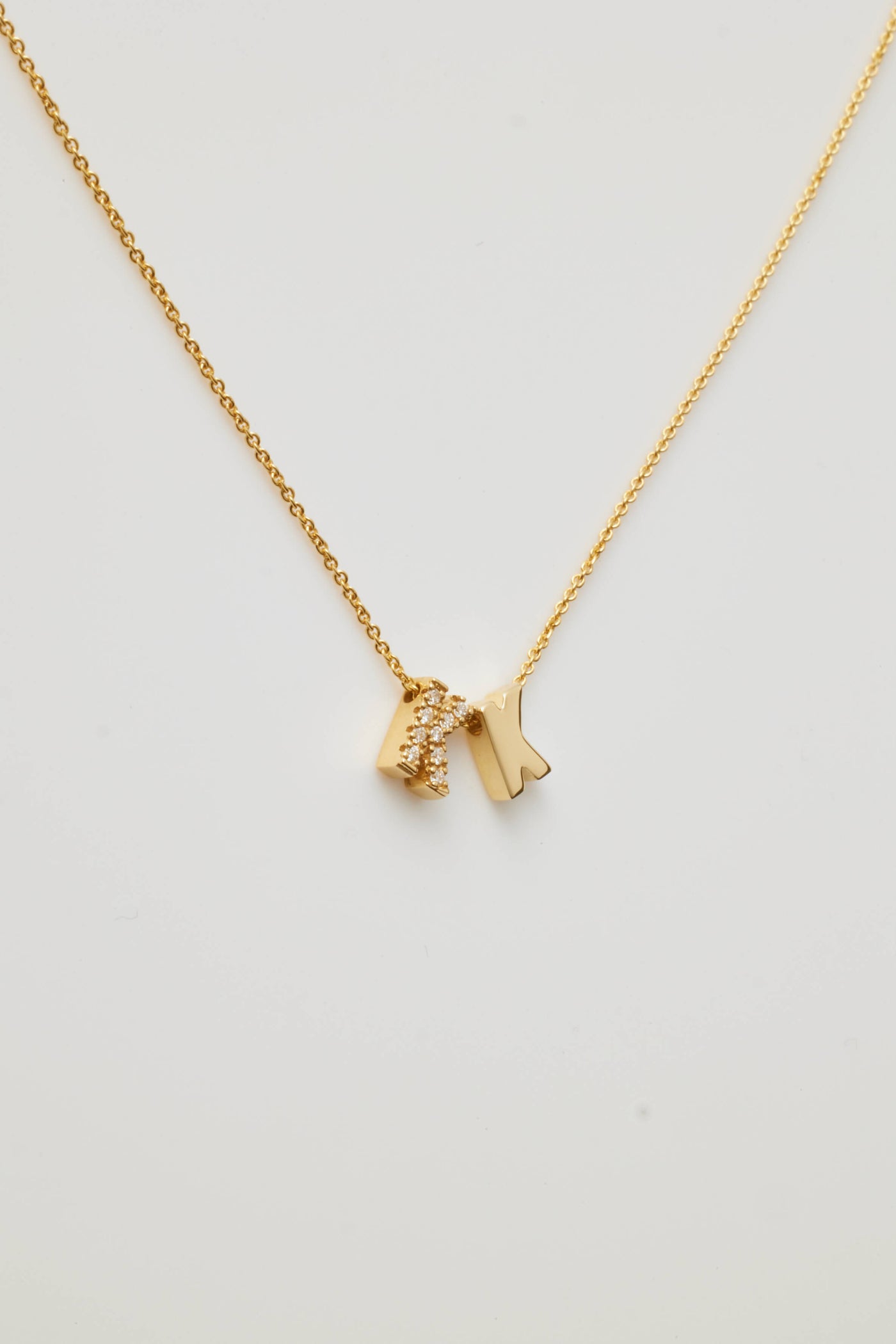 The Two Letters Necklace in Diamonds and 18k Gold showcases a minimalist and elegant design with two hanging pendants: a solid 18k gold "K" and a gold "K" encrusted with sparkling diamonds. This stunning necklace shines beautifully against a grey background.