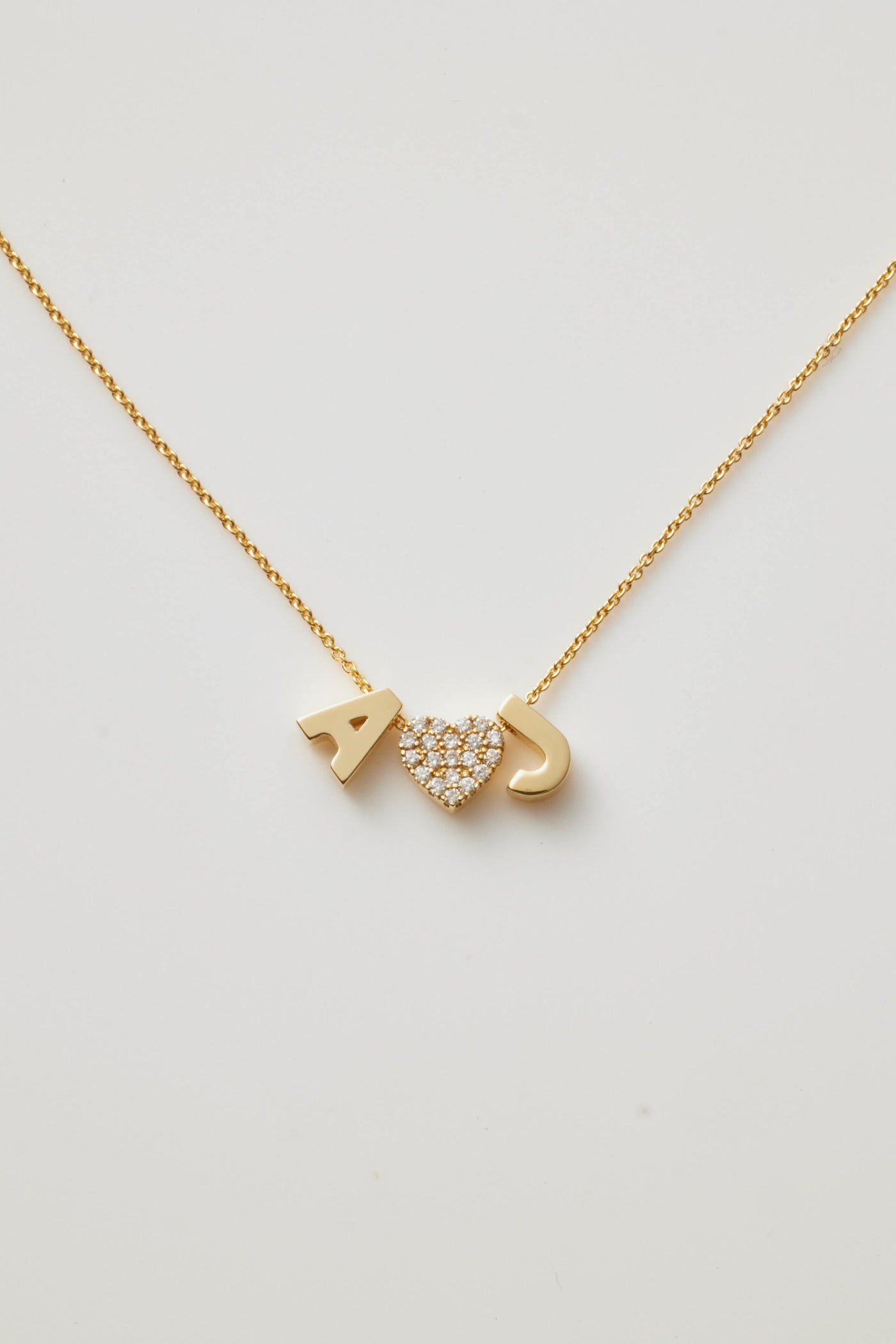 Introducing the Two Letters and Diamond Heart Necklace in 18k Gold: a delicate, solid gold piece that beautifully features the letters "A" and "J" alongside a heart-shaped charm embellished with small, sparkling diamonds nestled between them. Its thin and elegant chain flawlessly complements the minimalist design.