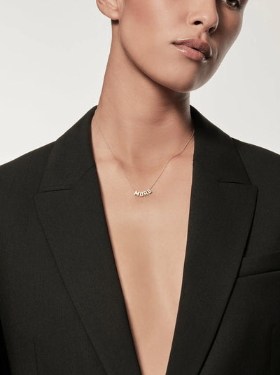 A person is wearing a personalized letter necklace letters "MBRB"  crafted in 18k solid gold, paired with a tailored black blazer with an open front that reveals bare skin underneath. The image prominently focuses on the Four Letters Necklace and the upper part of the blazer.