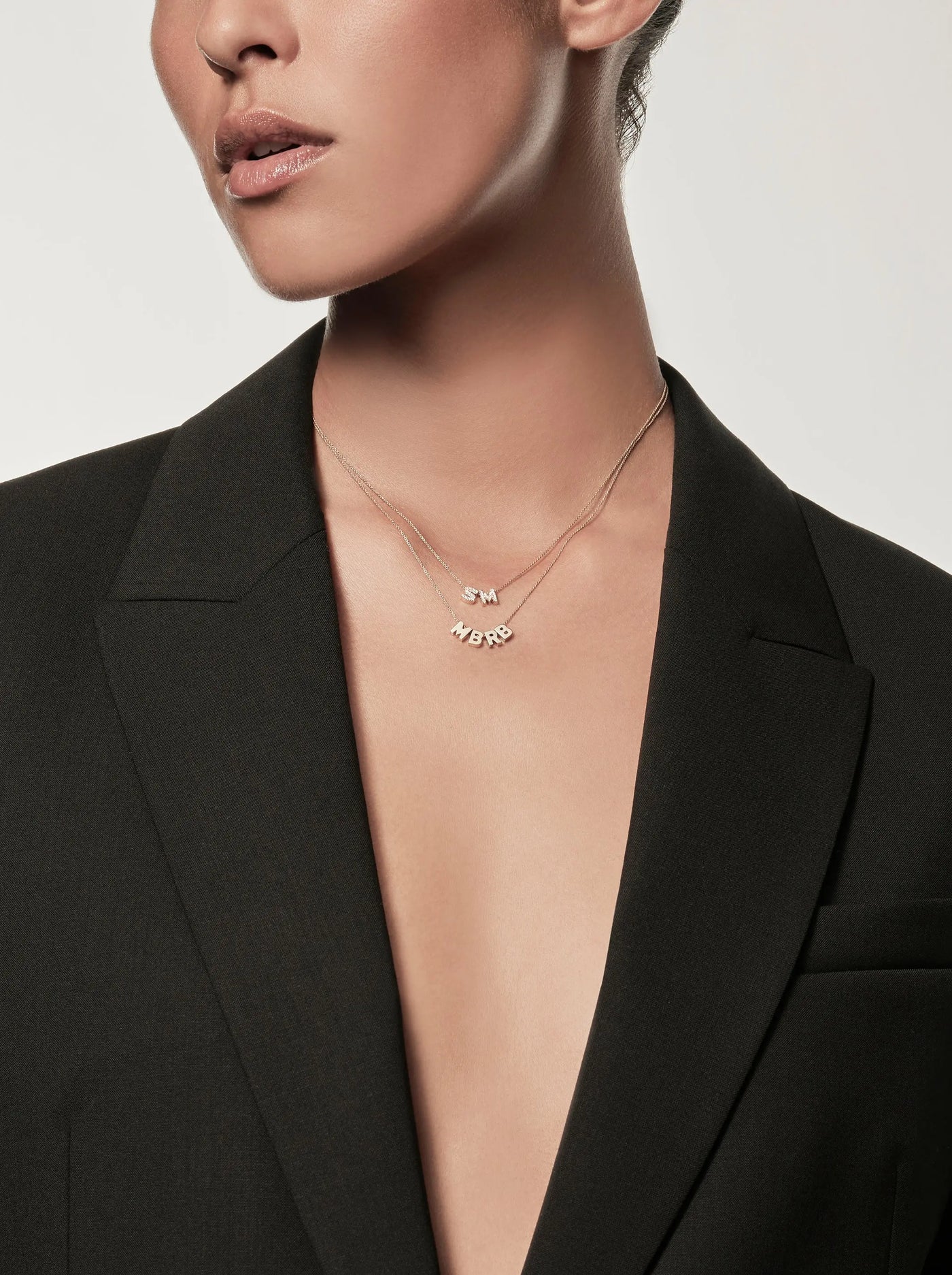 A person wearing a dark blazer with an open neckline and two charms slides onto two necklaces. Four Letters Necklace in 18k Gold initials "SM," while the other initials are "MBRB" The background is plain and light-colored, with the person's face partially visible, focusing on the jawline and lips.