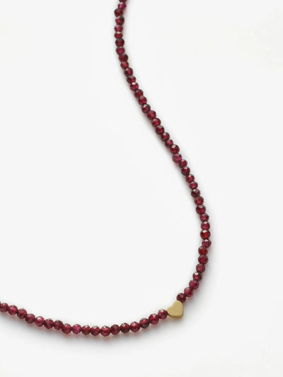 A person is shown from the neck down, donning a deep V-neck blazer over a bare chest. They're accessorized with the Heart Necklace in Garnet and 18k Gold, adding an elegant touch to their ensemble. The plain light background accentuates the minimalistic and sophisticated fashion style.