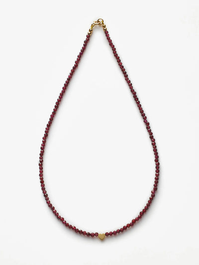 A person is shown from the neck down, donning a deep V-neck blazer over a bare chest. They're accessorized with the Heart Necklace in Garnet and 18k Gold, adding an elegant touch to their ensemble. The plain light background accentuates the minimalistic and sophisticated fashion style.