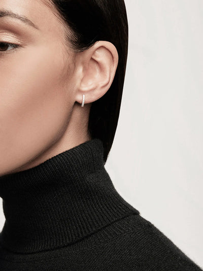 A close-up image of a person wearing a black turtleneck sweater, with their face partially visible. They have short, dark hair and a small Huggie Earring in Diamonds and 18k Gold in their ear. The background is plain and light-colored, highlighting the minimalistic and elegant style.