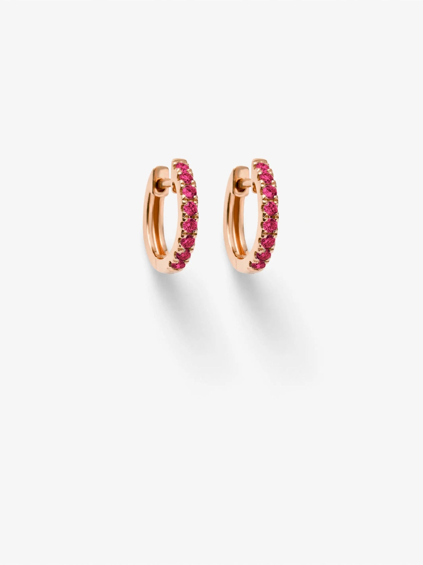 A pair of Huggie Earrings in Ruby and 18k Rose Gold is displayed against a white background. The elegant smaller hoops, encrusted with vibrant pink gemstones, are positioned symmetrically, casting faint shadows beneath them.