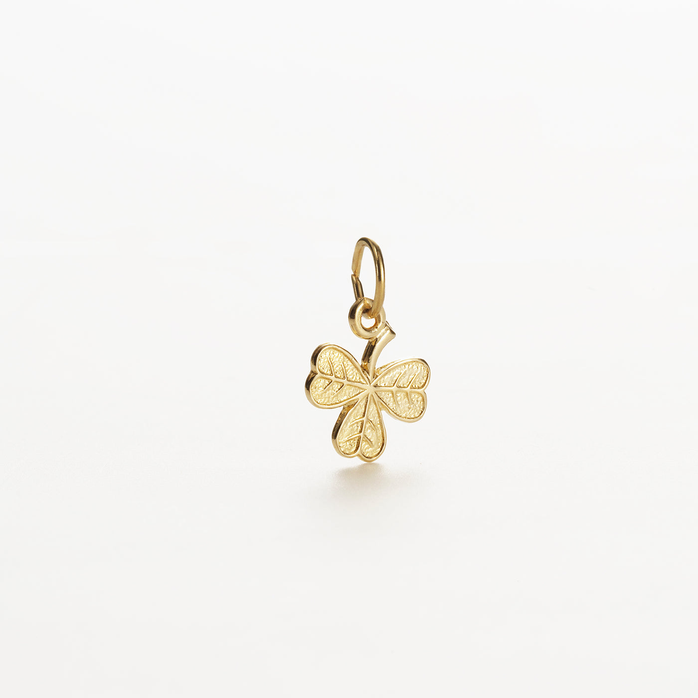 The Shamrock Charm in 9k Gold, measuring 12 mm, hangs against a plain white background. The clover leaves are intricately engraved with delicate details, and it is fitted with a small loop for attachment to a chain or bracelet.