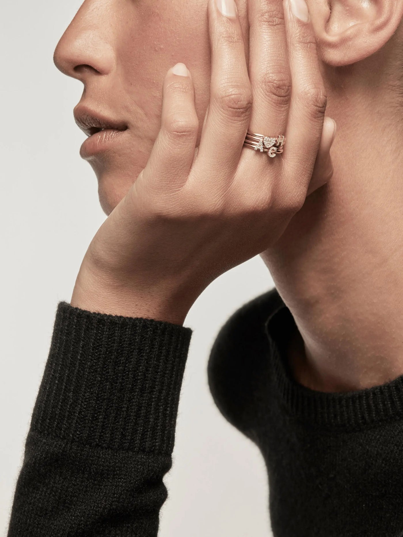 Close-up of a person resting their chin on their hand, showing a side profile of their face. They are wearing a black sweater and several rings, including the Letter C Ring in Diamonds and 18k Gold, on their fingers. The background is plain and light-colored.