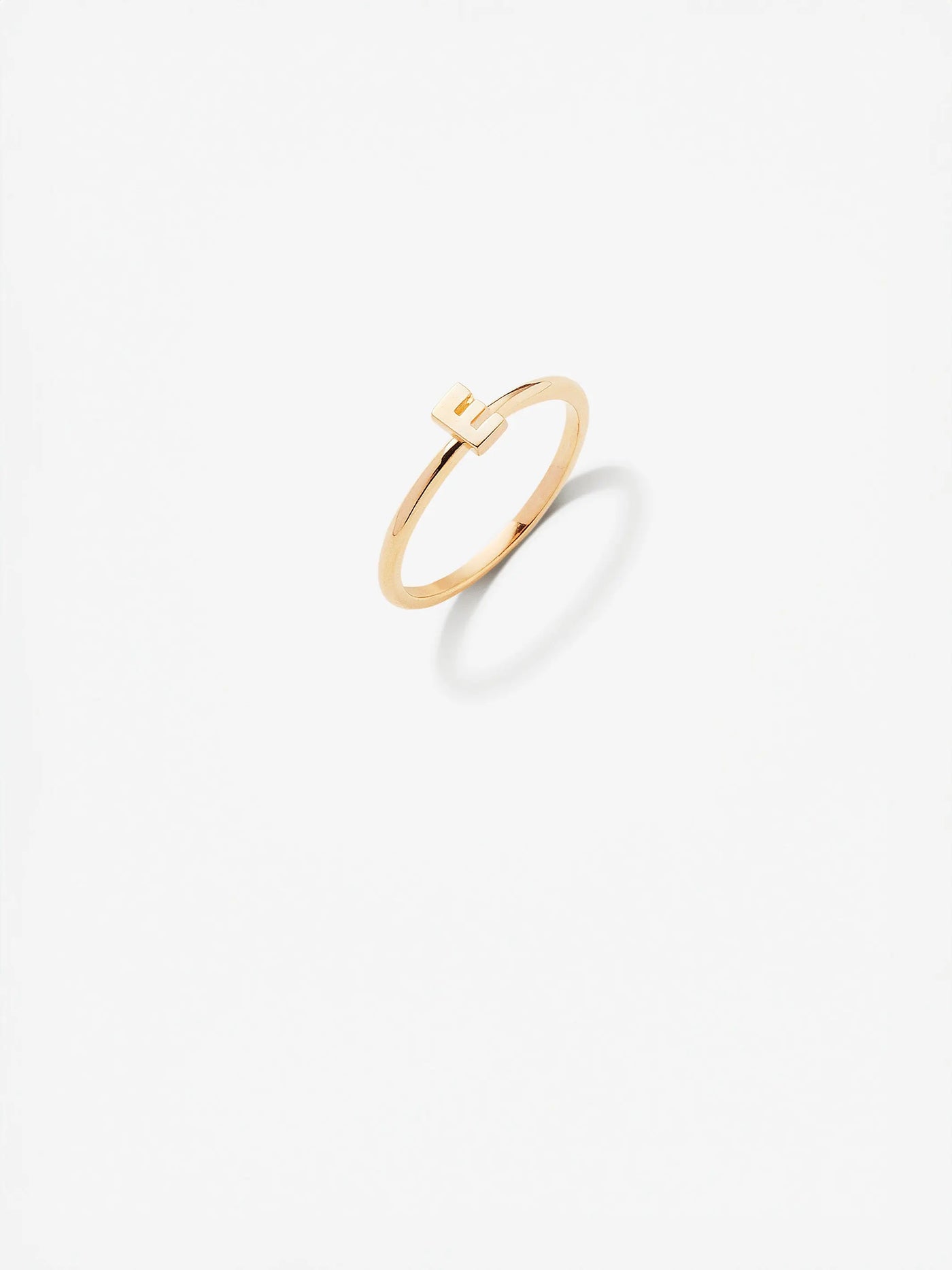 The Letter E Ring in 18k Gold is a delicate piece with a minimalistic design, featuring a small, cut-out square detail at the top center. Displayed against a plain white background, the ring casts a faint shadow that adds depth to the image. Available in custom sizes for the perfect fit.