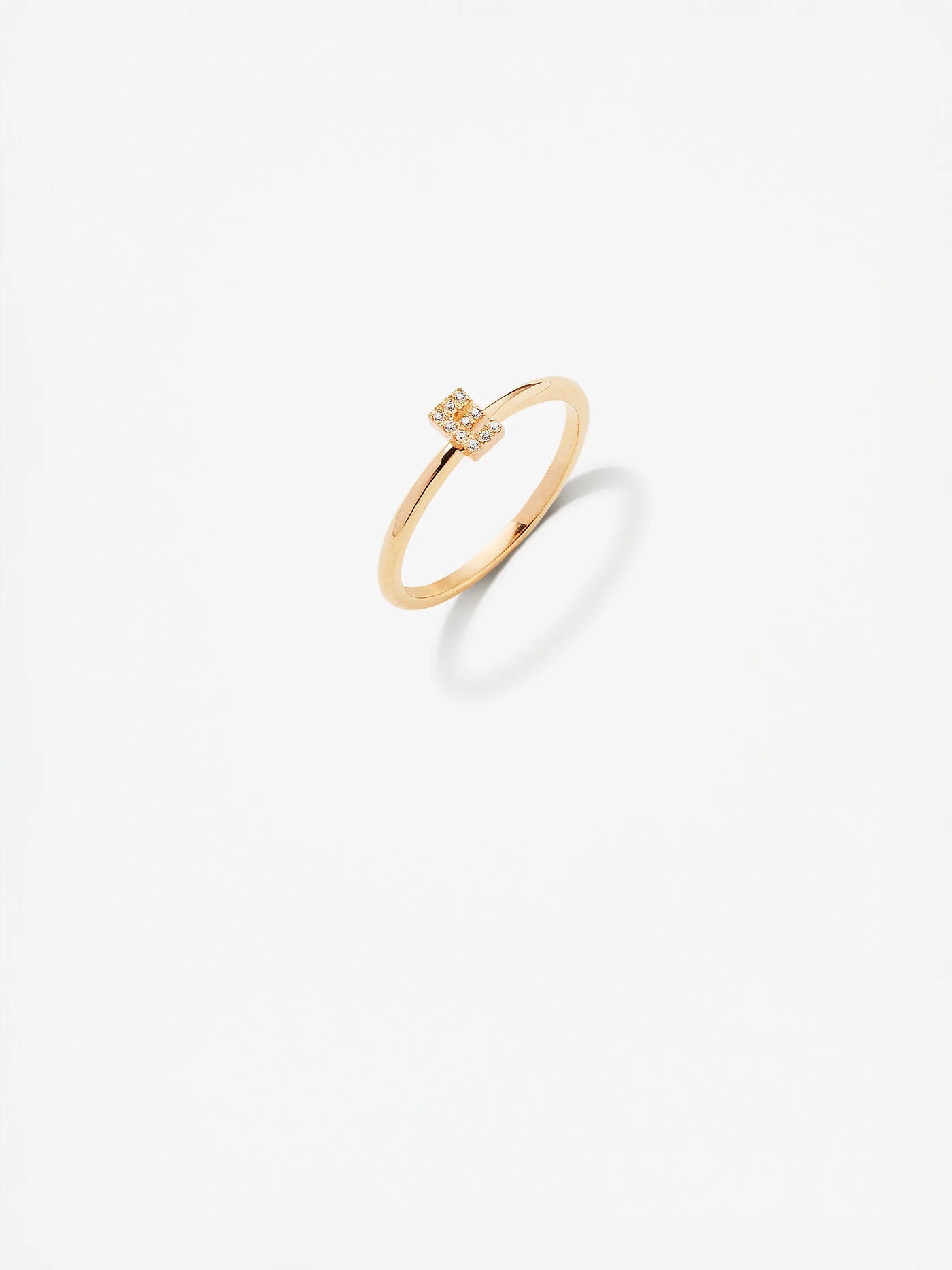 An elegant 18k gold Letter E Ring in Diamonds, featuring a minimalist design with a small square setting encrusted with tiny diamonds. Displayed on a white background, the ring casts a delicate shadow beneath it.