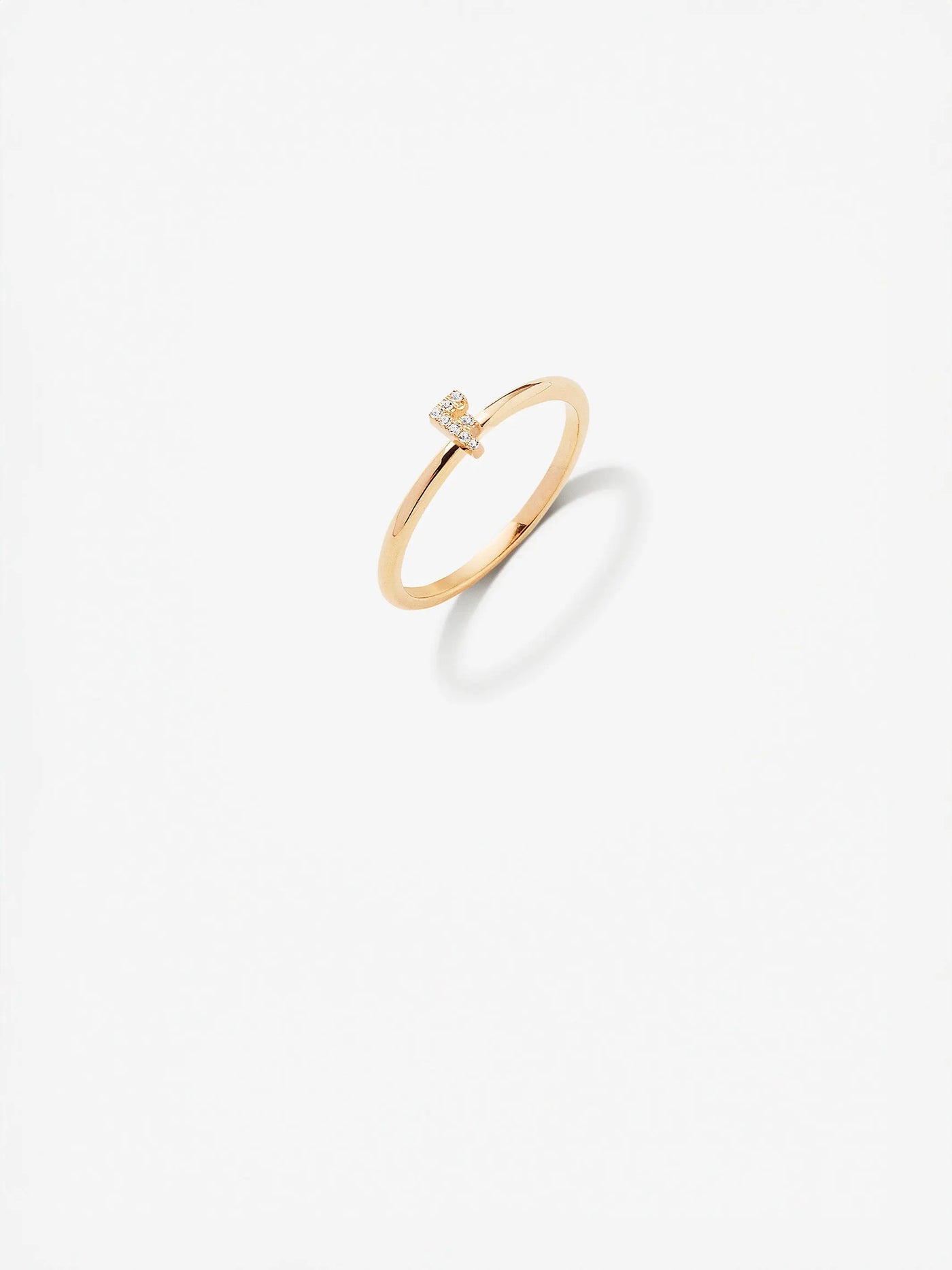 The "Letter F Ring in Diamond and 18k Gold," featuring a small diamond setting, is centered on a white background. The ring's simple and elegant design casts a subtle shadow beneath it.