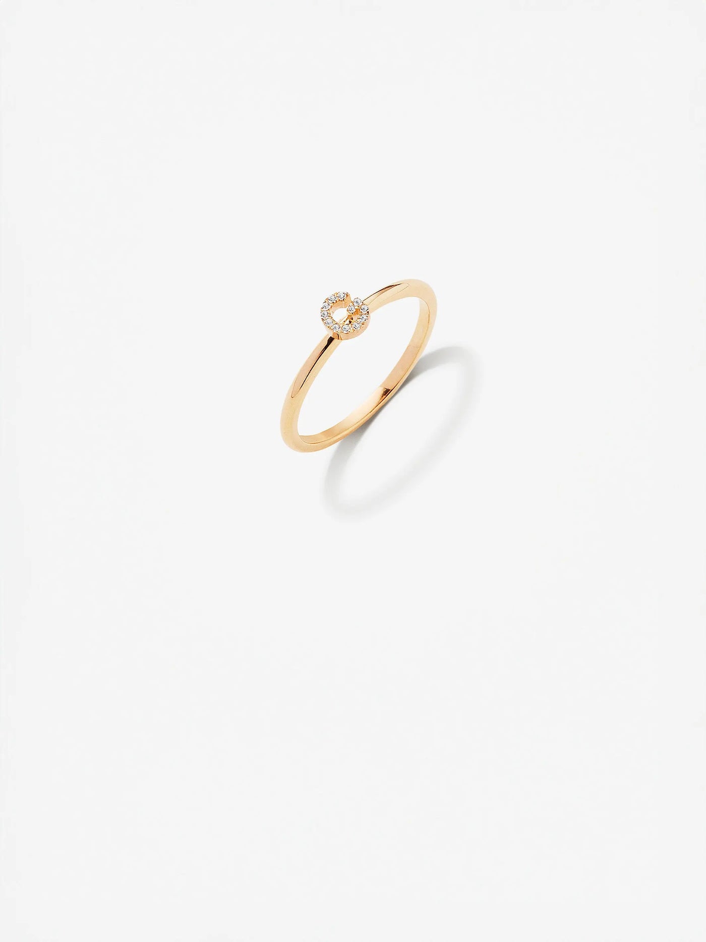 The Letter G Ring in Diamonds and 18k Gold, featuring a delicate design with a small circular setting adorned with brilliant diamonds, is displayed against a white background. The ring casts a slight shadow below, adding depth to the simple and elegant presentation.