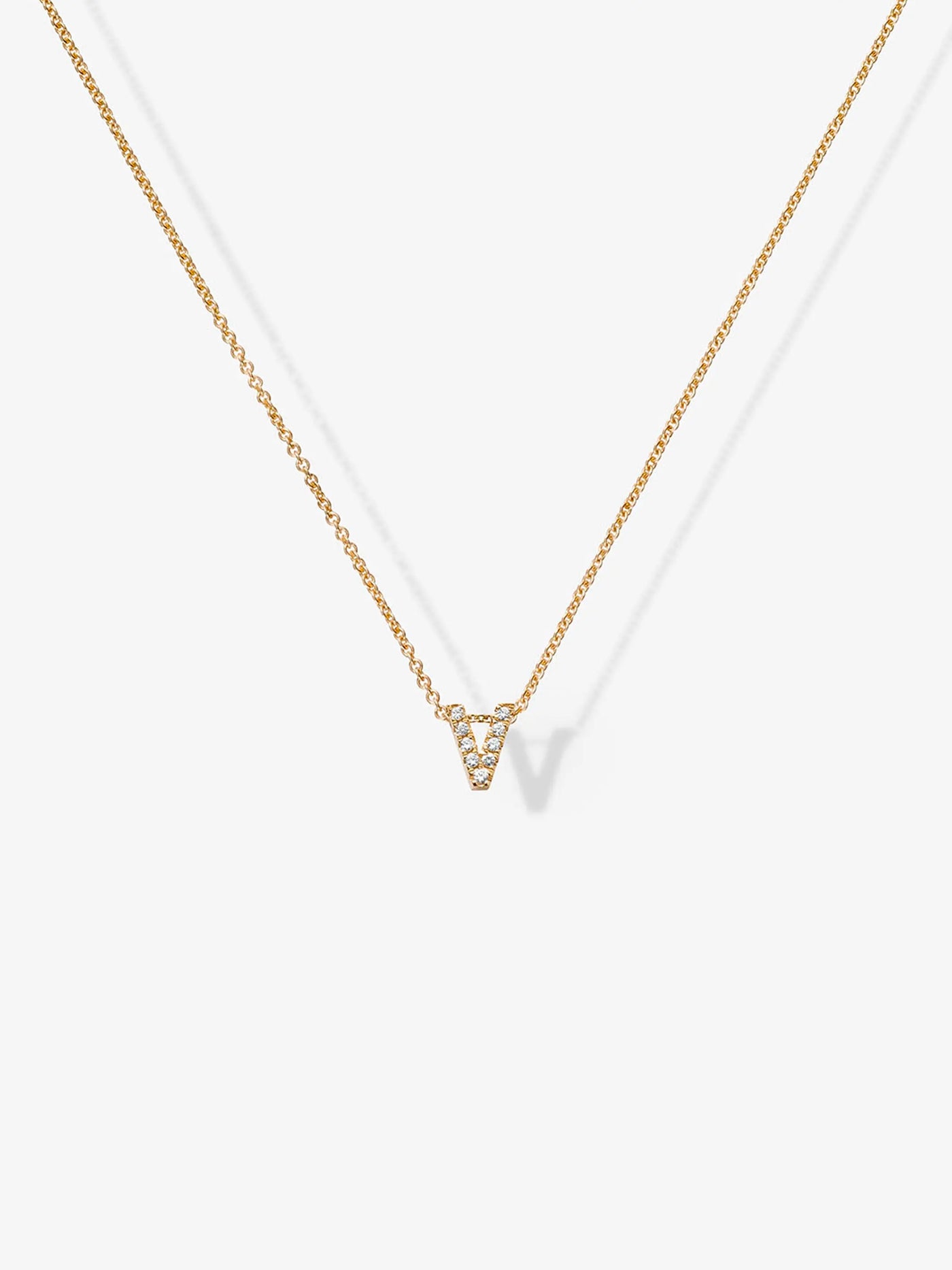 A delicate "Letter V Necklace in Diamonds and 18k Gold," featuring a thin chain and a small, V-shaped pendant encrusted with tiny, sparkling diamonds, is photographed against a plain white background. The necklace's shadow is visible.