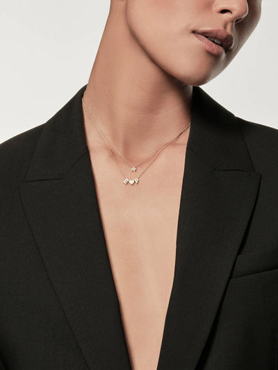 A person is wearing a black blazer without a shirt underneath, adorned with layered gold chains featuring small pendants. The focus is on the neck and upper chest, with the person's face partially visible. One of the pendants is the One Diamond Star Necklace in 18k Gold, sparkling subtly against the plain, light-colored background.