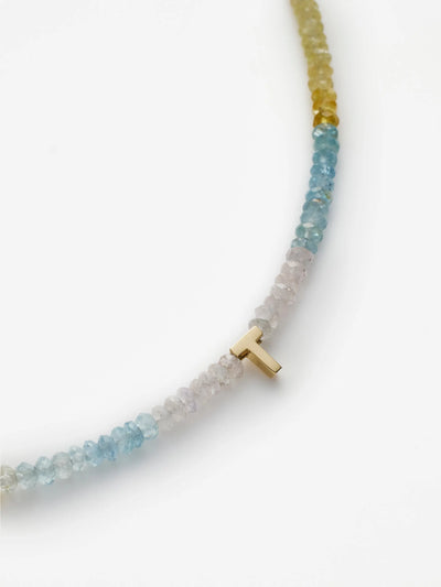 A close-up image of the One Letter Bracelet in Aquamarine and 18k Gold, featuring a small 18k solid gold 'T' charm. The beads, reminiscent of an aquamarine bracelet, vary in color from white to shades of light blue and yellow, creating a gradient effect that evokes mental clarity. The bracelet is displayed on a plain white background.