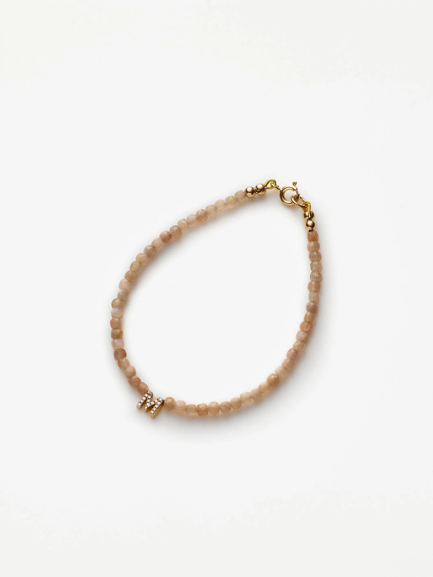 The One Letter Bracelet in Diamonds, Blush Moonstone and 18k Gold showcases a delicate arrangement of small, round blush pink moonstone beads. It features an 18k solid gold clasp and a tiny gold accent embedded with clear crystals, all beautifully displayed in a circular shape on a plain white background.