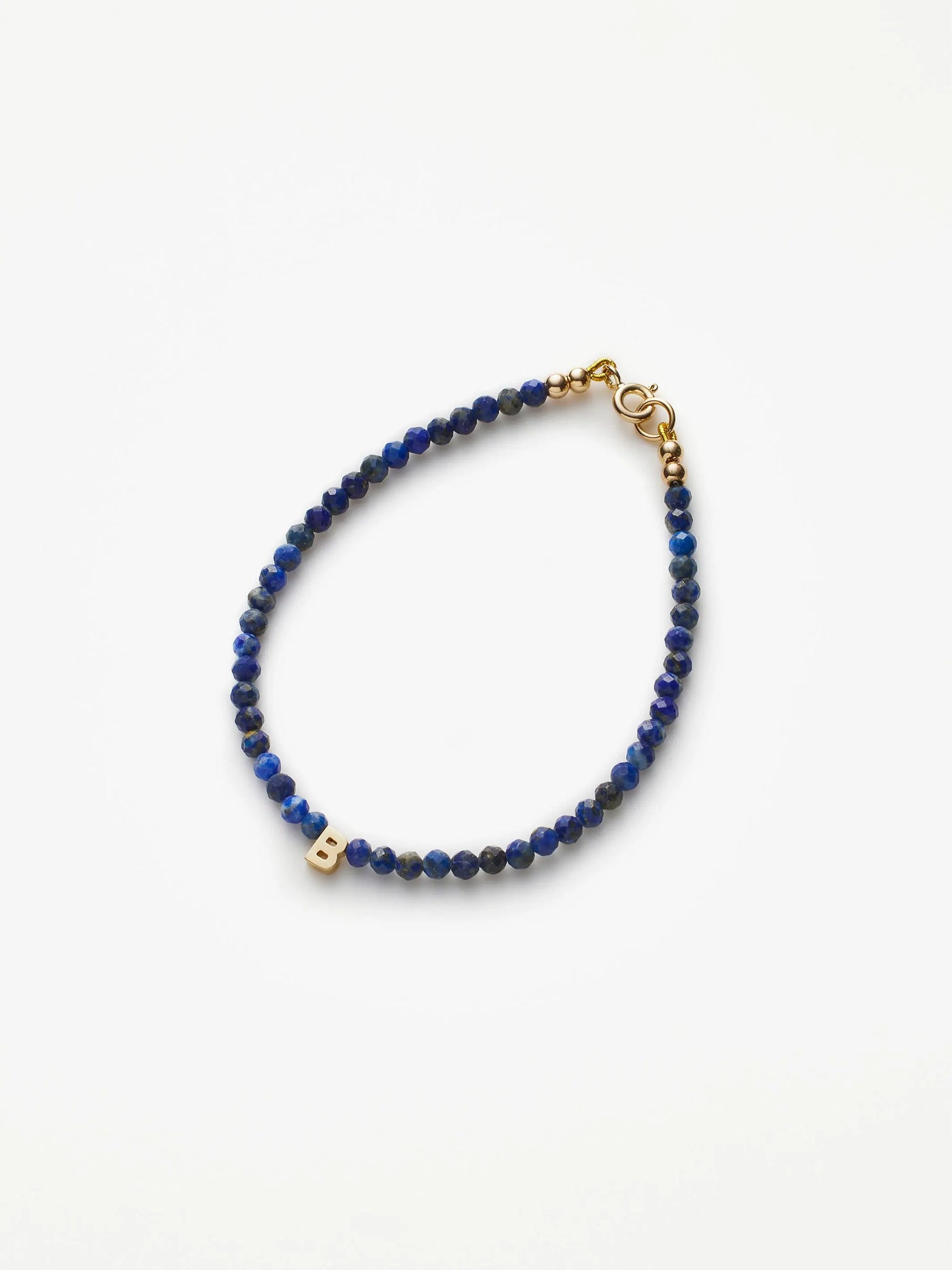 The One Letter Bracelet in Lapis Lazuli and 18k Gold is a delicate piece featuring small, round lapis lazuli beads and an 18k gold clasp. Among the hand-strung blue beads, a gold letter "B" charm is prominently placed, adding a personalized touch to this elegant bracelet, which is displayed on a plain white background.