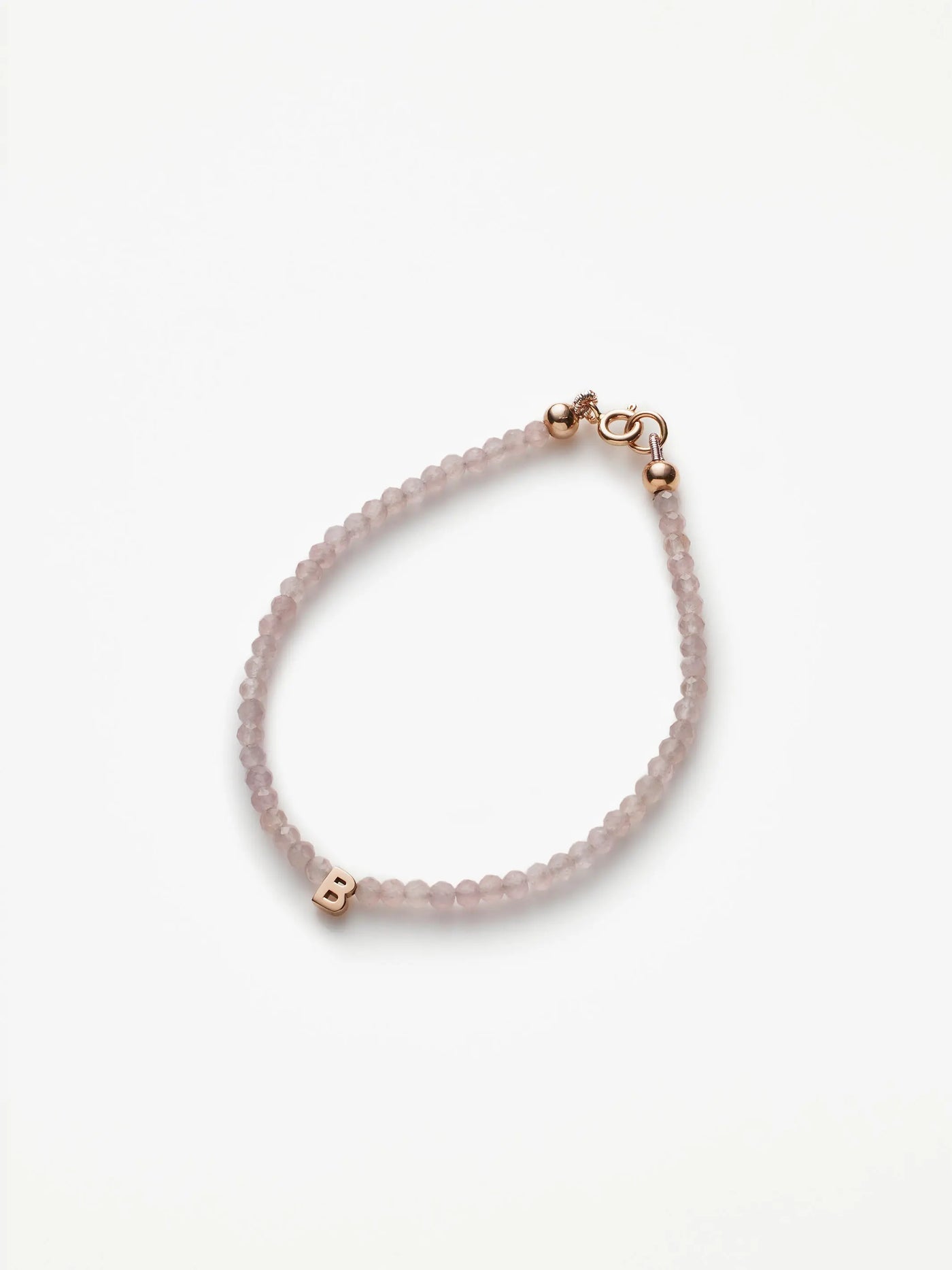 The One Letter Bracelet in Rose Quartz and 18k Rose Gold features small, round rose quartz beads and a gold lobster clasp. A single 18k gold initial charm, the letter "B," is threaded onto the beads. The bracelet is available in custom sizes, arranged in a simple, curved shape on a white background.