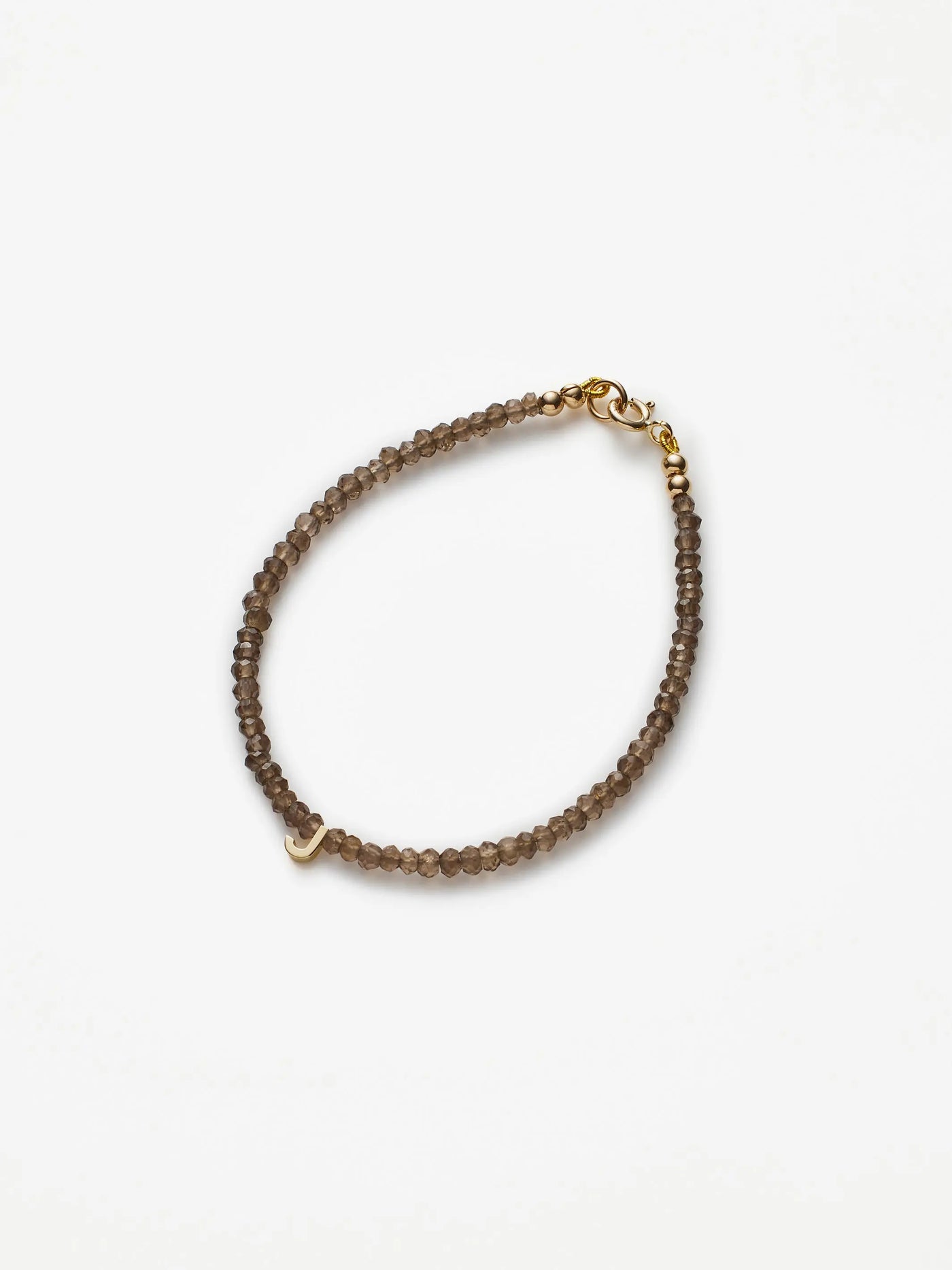 The One Letter Bracelet in Smoky Quartz and 18k Gold is crafted from small, round, translucent brown beads. It features an 18k solid gold clasp and a small gold heart charm. Shown arranged in an oval shape on a plain white background, custom sizes are available upon request.