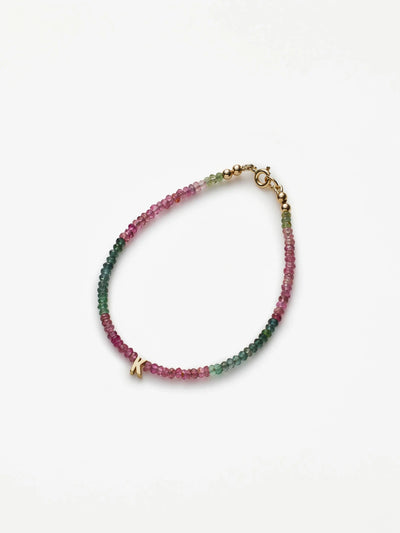 The One Letter Bracelet in Tourmaline and 18k Gold showcases delicate, small multicolored beads arranged in a beautiful gradient from pink to green. The piece is elegantly finished with an 18k solid gold clasp and a charming letter "K" charm. Displayed on a white background in an oval shape, the bracelet is available in custom sizes.