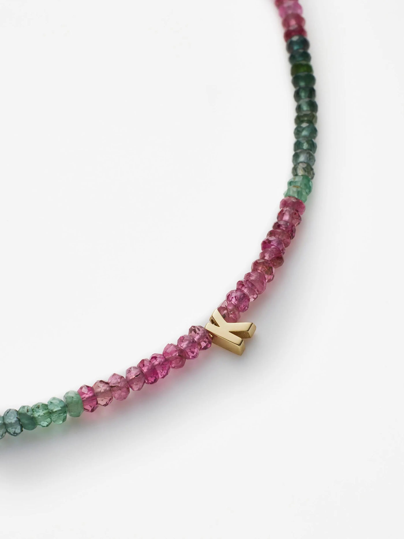 A close-up image of the "One Letter Bracelet in Tourmaline and 18k Gold" showcases small, multi-colored gemstones transitioning from green to pink. Among the vibrant beads, a small charm with an 18k solid gold letter "K" is attached, creating a striking contrast against the colorful gemstones. The background is plain white.