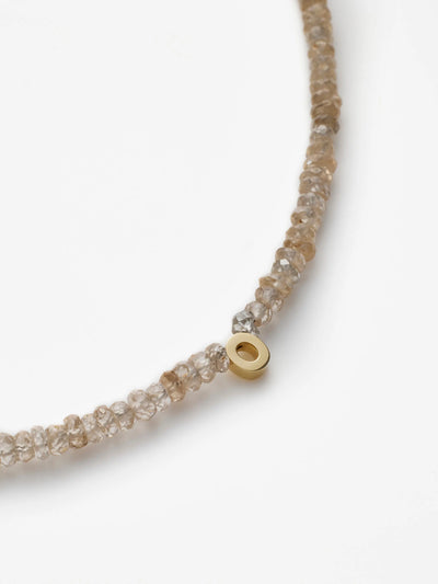 A close-up image of the One Letter Bracelet in Zircon and 18k Gold showcases its delicate craftsmanship, featuring translucent, faceted taupe zircon stones strung together. Among these elegant beads, a single small cylindrical bead made of 18k solid gold stands out prominently. The plain white background accentuates the finely crafted piece, which is available in custom sizes.