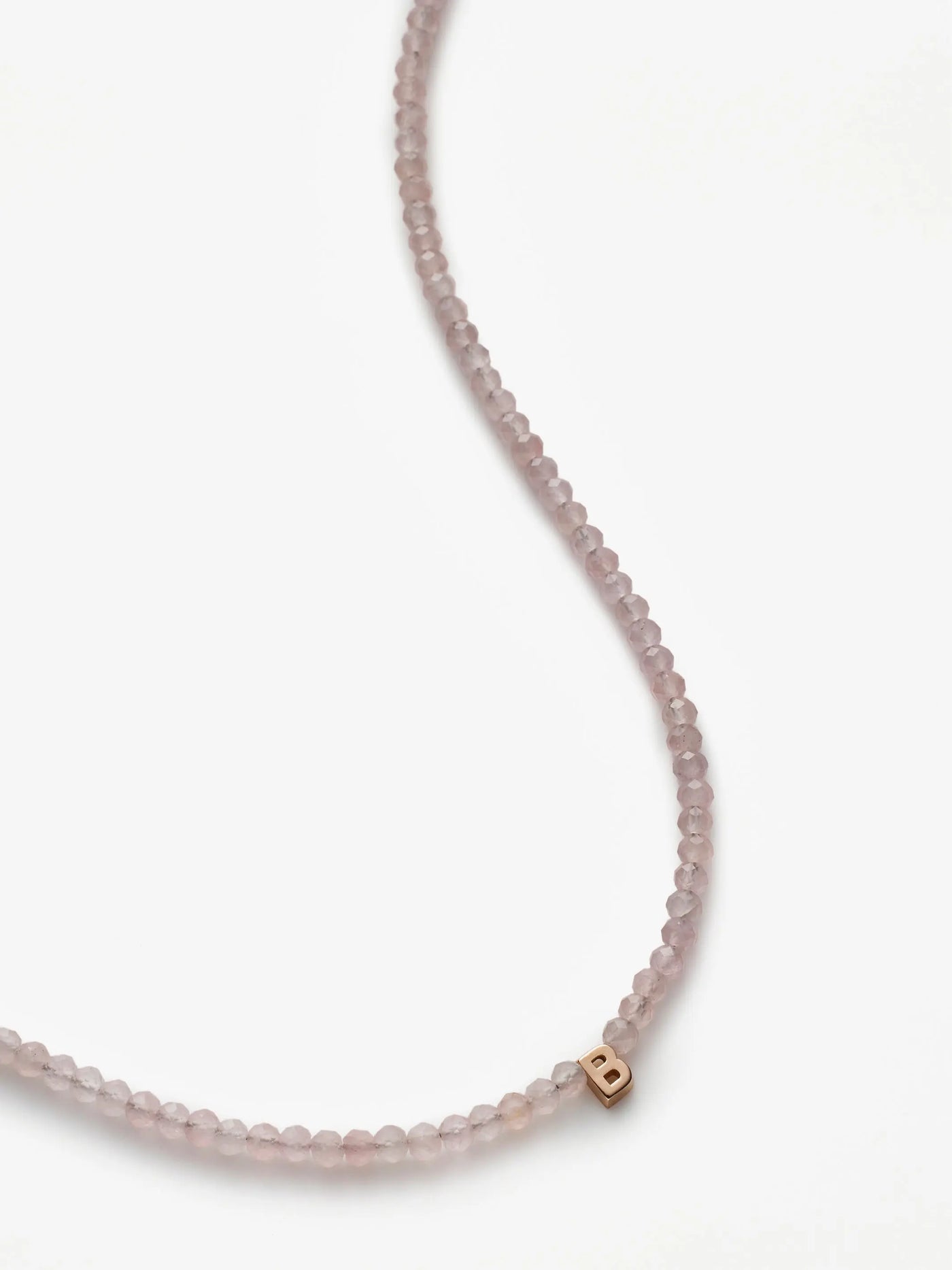 A delicate pink beaded One Letter Necklace in Rose Quartz and 18k Rose Gold with a subtle sheen is pictured against a white background. The necklace features a small cube-shaped 18k rose gold charm engraved with the letter "B" on one side.