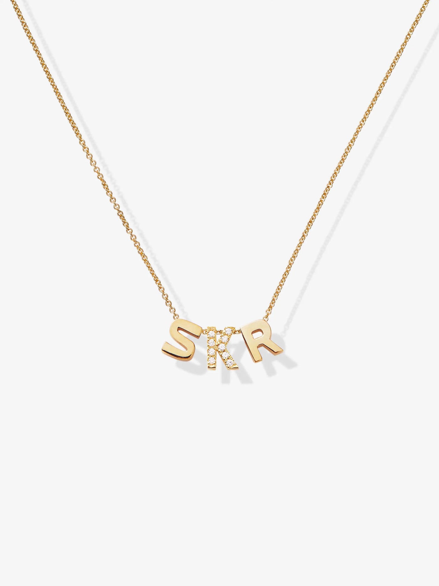 Introducing the Three Letters Necklace in Diamonds and 18k Gold: a stunning piece featuring a fine chain and three elegant letter charms. The "S" and "R" are crafted from solid 18k gold, while the "K" is encrusted with small, clear diamonds. Together, the letters spell out "SKR," evenly spaced along the chain. This exquisite necklace is beautifully showcased against a simple white background.