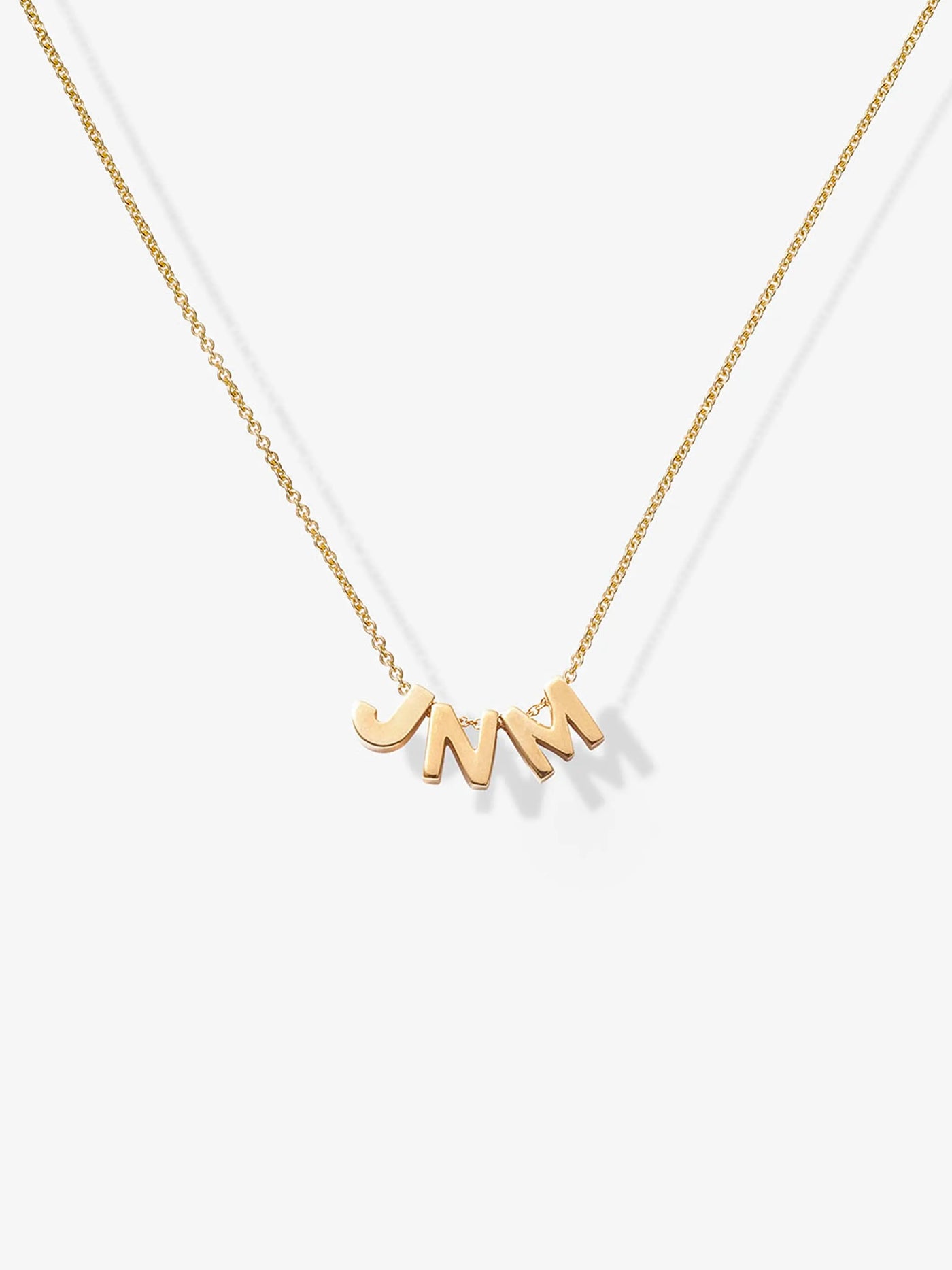 The Three Letters Necklace in 18k Gold is a delicate piece featuring three polished gold letters as the pendants. This sophisticated necklace, with its fine chain, gracefully displays the initials "JNM," which hang prominently at the center, casting a slight shadow on any light background.