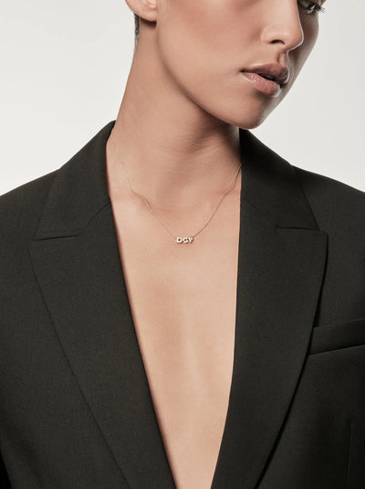 A person is shown from the shoulders up, wearing a black blazer with a deep neckline. Three Letters Necklace in 18k Gold, which features small charms spelling "DOP." The background is plain and light-colored.