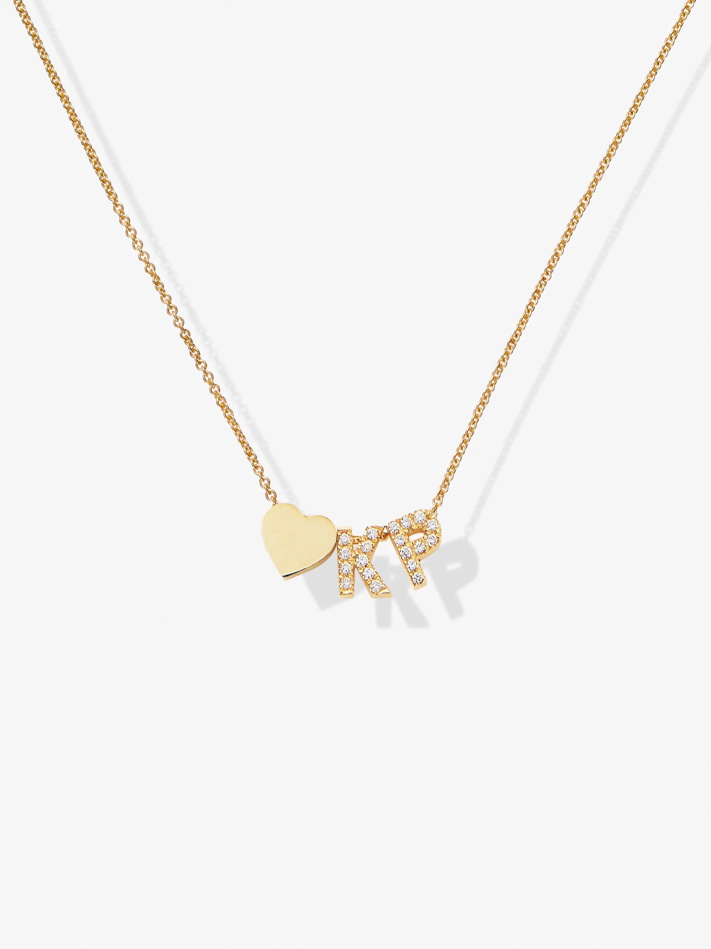 The "Two Letters and Heart in Diamonds and 18k Gold" is a delicate 18k solid gold necklace featuring a heart-shaped pendant adorned with the letters "MR" in small crystals. The thin gold chain includes an openable connector clasp for ease of wear. Displayed on a plain white surface, the heart and letters create an elegant presentation.