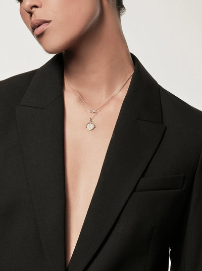 A person wearing a black blazer with no shirt underneath is shown from the shoulders down. They are also wearing the Two Letters Necklace in 18k Gold, delicately designed with one small charm and one round pendant. The background is plain and light-colored.