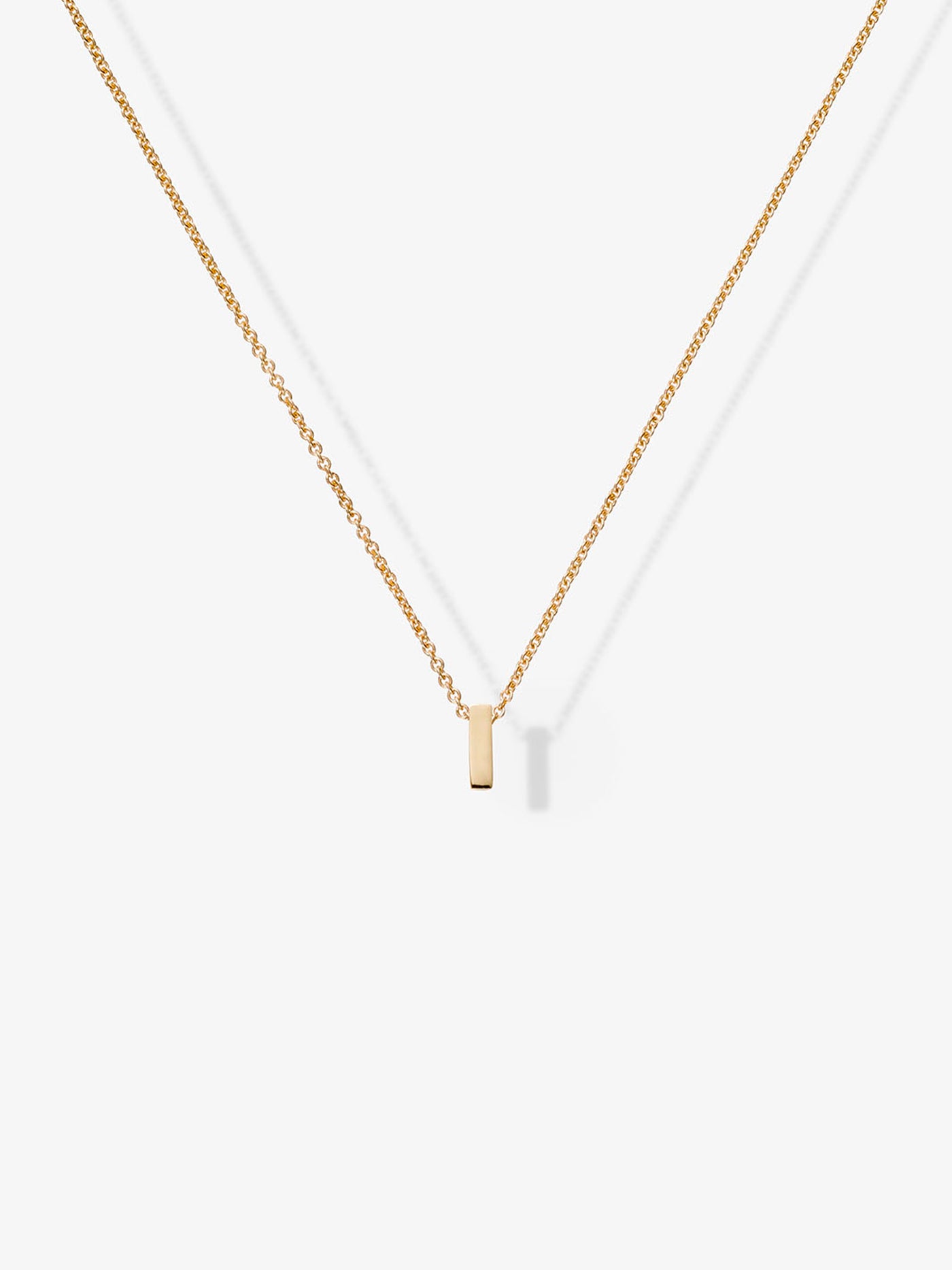 The Letter I Necklace in 18k solid gold features a small, rectangular gold pendant symmetrically centered on a delicate chain, casting a soft shadow on a white background.