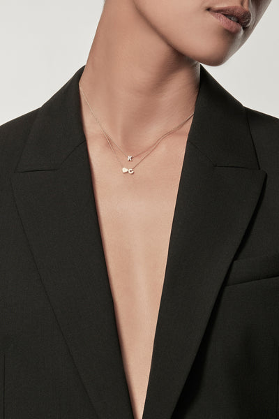 Close-up of a woman wearing a minimalist 18k gold initials “K”  and gold heart charm initial "C" necklace on a fine chain, styled with a black blazer – personalised alphabet pendant jewellery