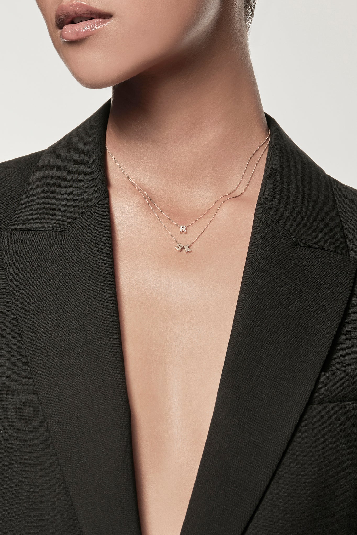 Close-up of a woman wearing a minimalist 18k gold initials “S” "K" necklace and diamond letter "R" necklace on a fine chain, styled with a black blazer – personalised alphabet pendant jewellery