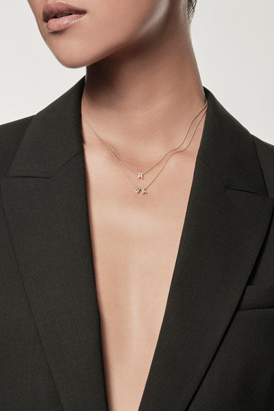 Close-up of a woman wearing a minimalist 18k gold initials “S” "K" necklace and diamond letter "R" necklace on a fine chain, styled with a black blazer – personalised alphabet pendant jewellery