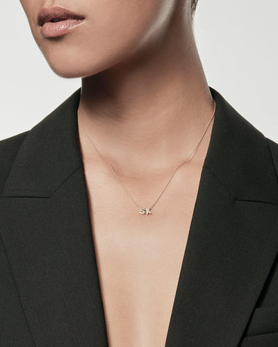 A person wearing a black blazer with no shirt underneath, showcasing a delicate Two Letters Necklace in 18k Gold with the initials "SK" at the center. The background is a plain light gray. The person's face is mostly out of frame, focusing on the jewelry and attire.