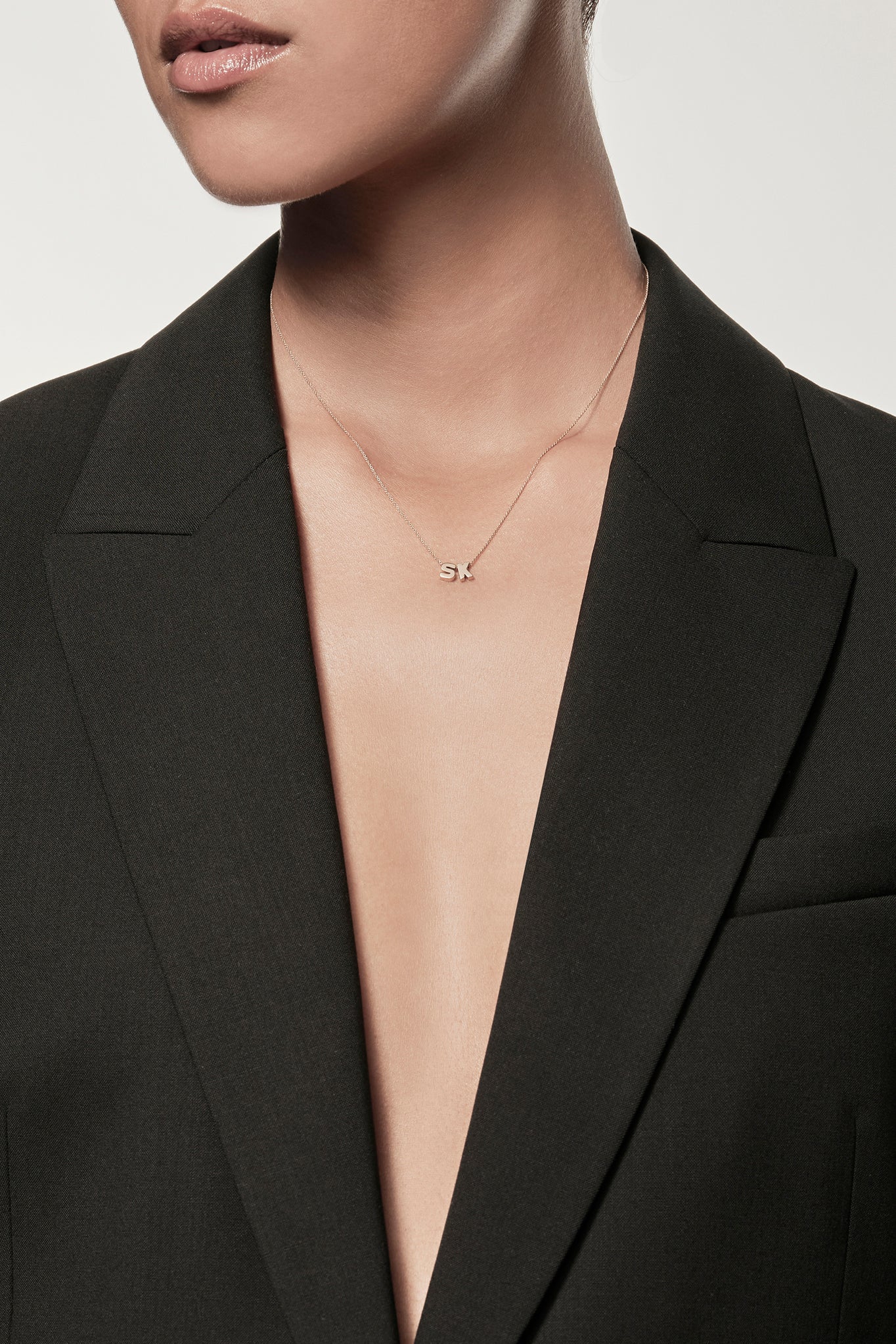 Close-up of a woman wearing a minimalist 18k gold initials “S” "K" necklace on a fine chain, styled with a black blazer – personalised alphabet pendant jewellery