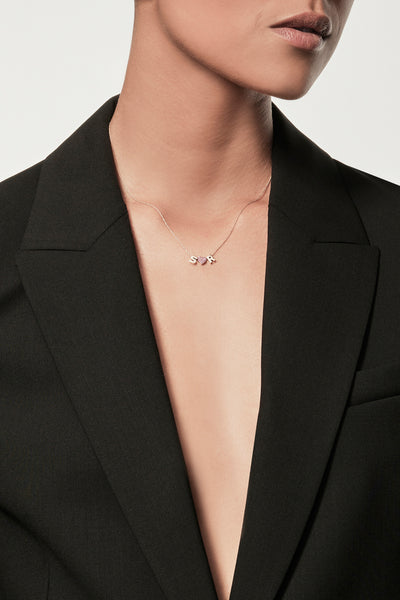 Close-up of a woman wearing a minimalist gold initial “S” and "R" separated with a pink sapphire heart charm, styled with a black blazer – personalised alphabet pendant jewellery