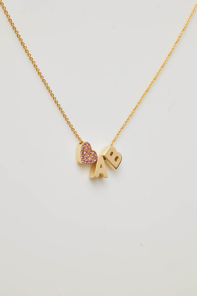 Gold necklace with two letter charms and a pink sapphire heart charm, symbolising love and personal significance.