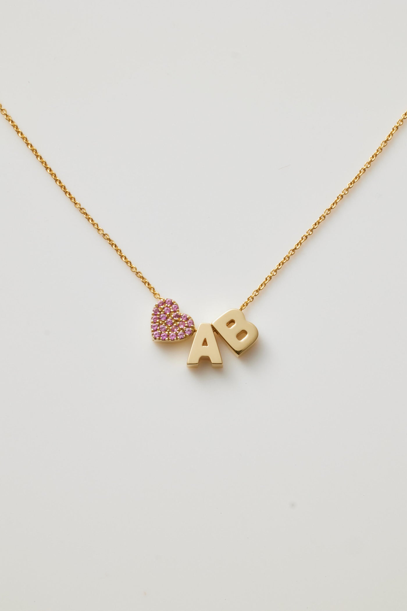 Gold necklace with two letter charms and a pink sapphire heart charm, symbolising love and personal significance.