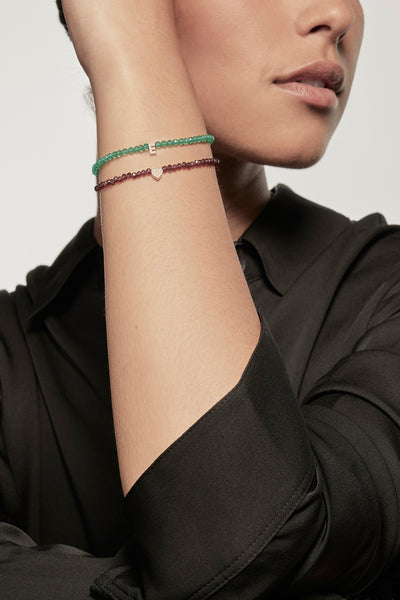 A faceted garnet petite bracelet with a gold and diamond heart charm is worn on the left wrist of a model dressed in a black silk shirt. The bracelet's deep red garnets contrast with the shirt's dark fabric, and the small gold heart adds a delicate accent.