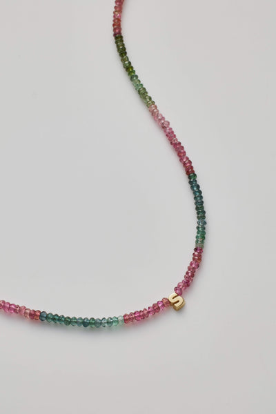 The One Letter Necklace in Tourmaline and 14k Gold showcases a delicate design with small, multicoloured tourmaline gemstones in green, pink, and red shades. It includes a slightly off-centre, small 14k gold 'S' charm. The beads are threaded on a fine, near-invisible string for a minimalist appearance.